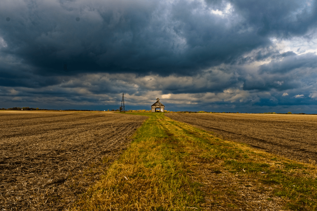 An image of a derelict farmhouse in the center of an open and recently harvested field that spans the bottom half of the frame. The farmhouse lies center frame with the upper half of the frame showing a dark cloudy sky.